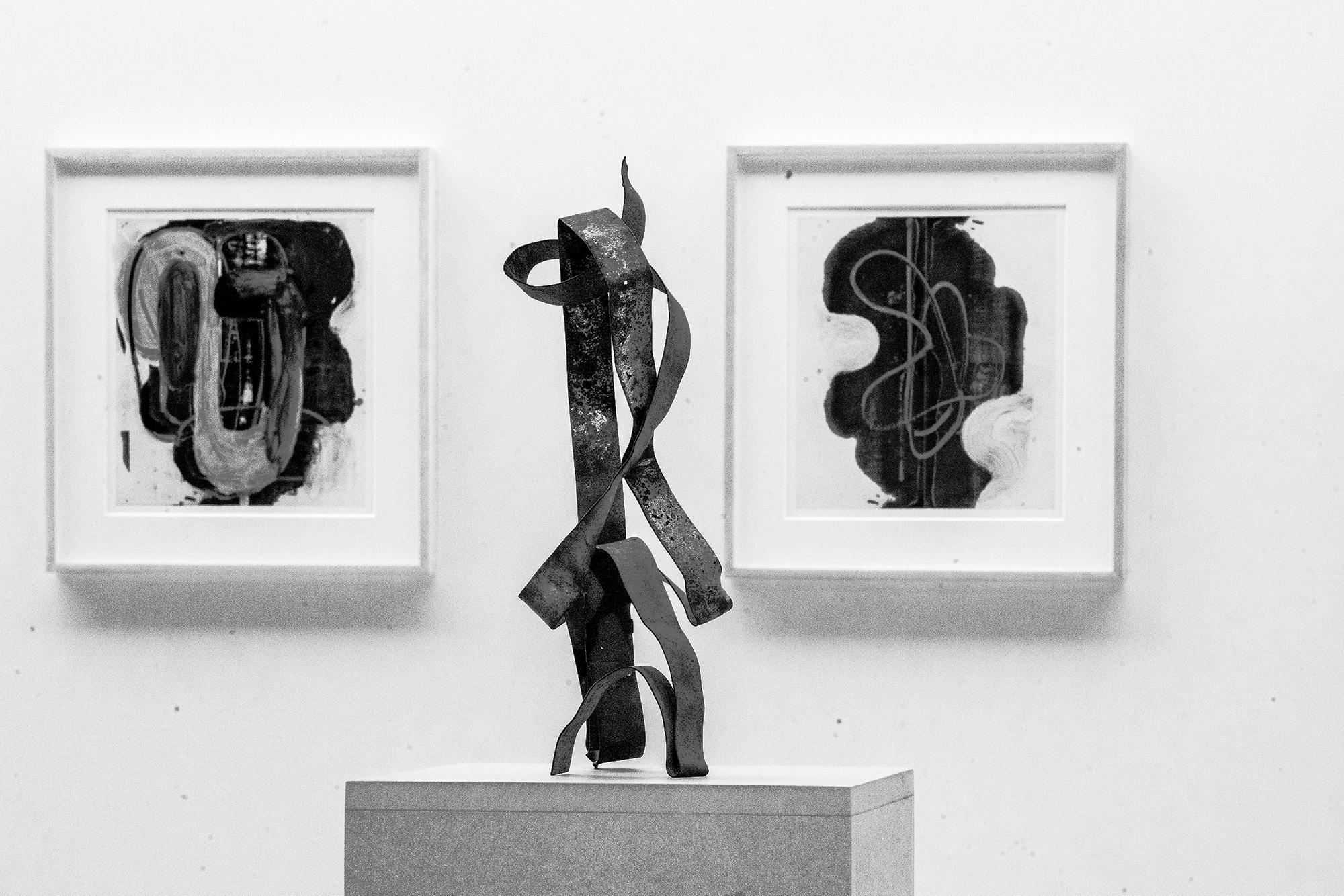 A black and white photograph documents three works of art. Two small framed abstract works on paper hang on the wall behind a small sculpture, consisting of bent metal, stands on a pedestal.