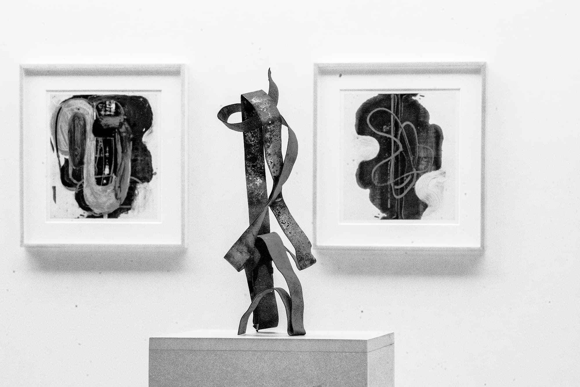 A black and white photograph documents three works of art. Two small framed abstract works on paper hang on the wall behind a small sculpture, consisting of bent metal, stands on a pedestal.