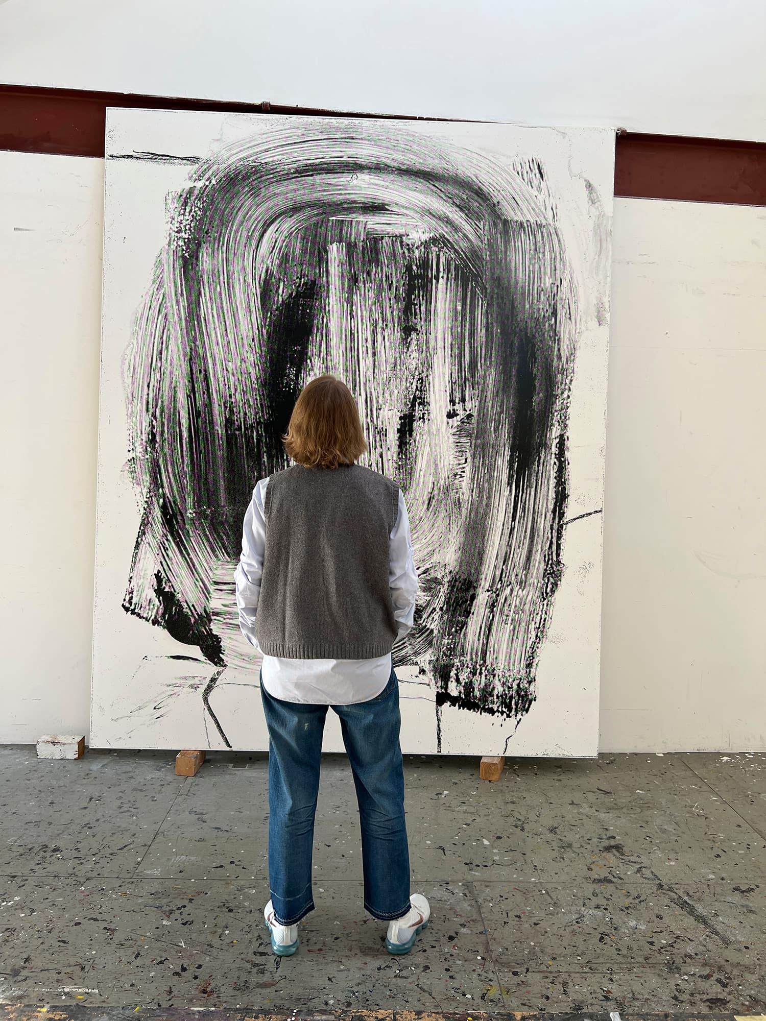 A color image of a person standing in front of a painting which leans on a wall. The person's back faces the camera. The painting appears to be enlarged brushwork, black pigment on a white support.